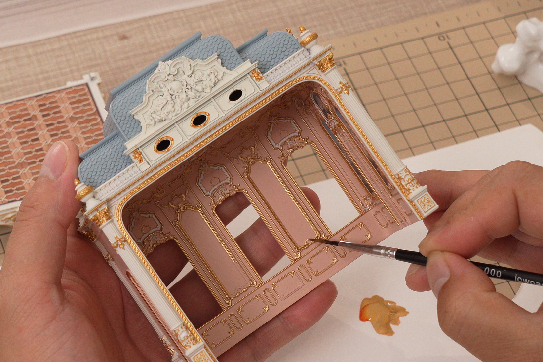 A fine paintbrush applying gold paint to the raised wall ornament of a miniature architectural interior